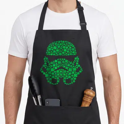 Clover st patrick day aprons - Green stormtrooper helmet shape formed by a cluster of small shamrocks — shown on a black cotton canvas apron against a white background