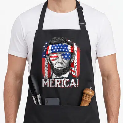 Abe 4th of july apron - Distressed Abraham Lincoln face wearing flag headband and sunglasses with MERICA text below — shown on a black cotton canvas apron against a white background