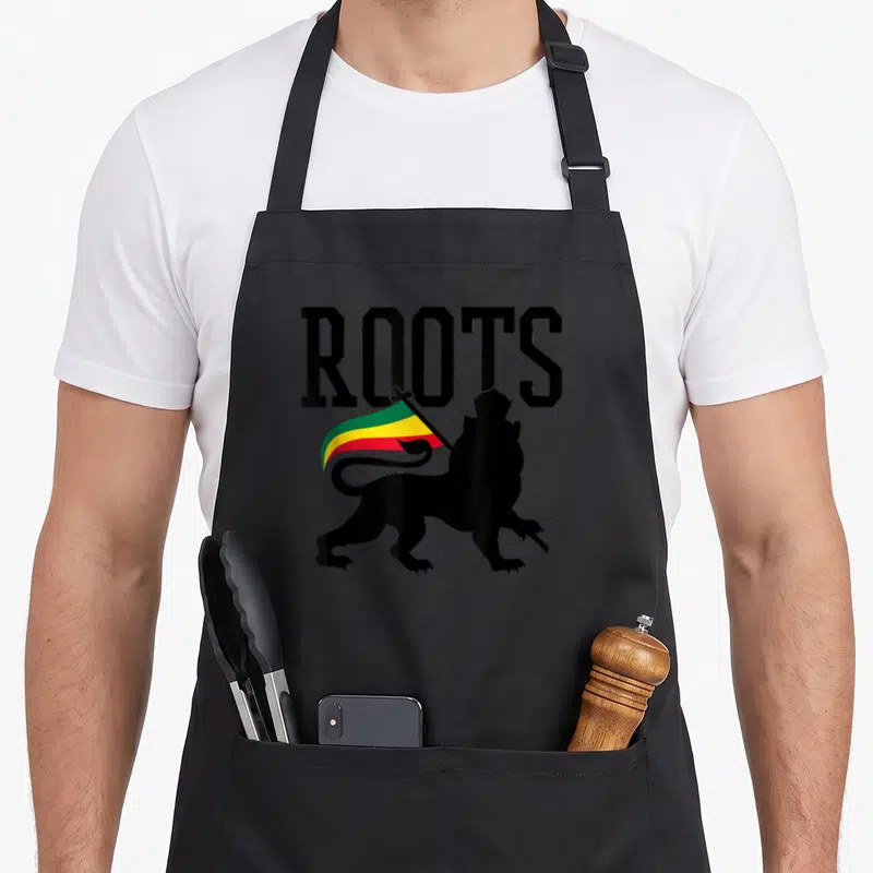 Reggae lions apron - ROOTS text above black lion silhouette holding green yellow red striped flag — shown on a black cotton canvas apron against a white background