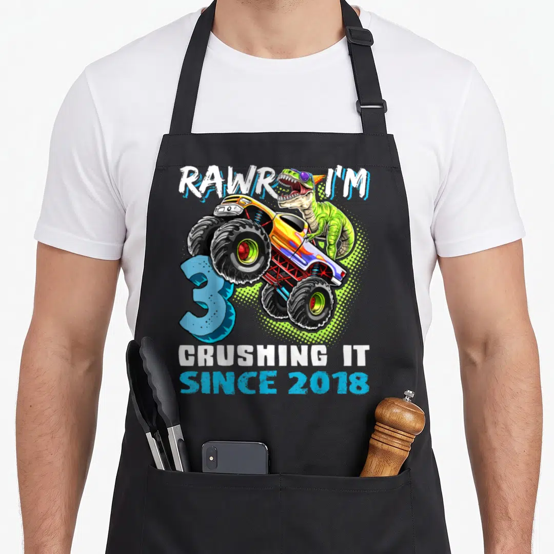 Rawr crushing truck apron - Green dinosaur driving yellow monster truck with 3 and 2018 text — shown on a black cotton canvas apron against a white background