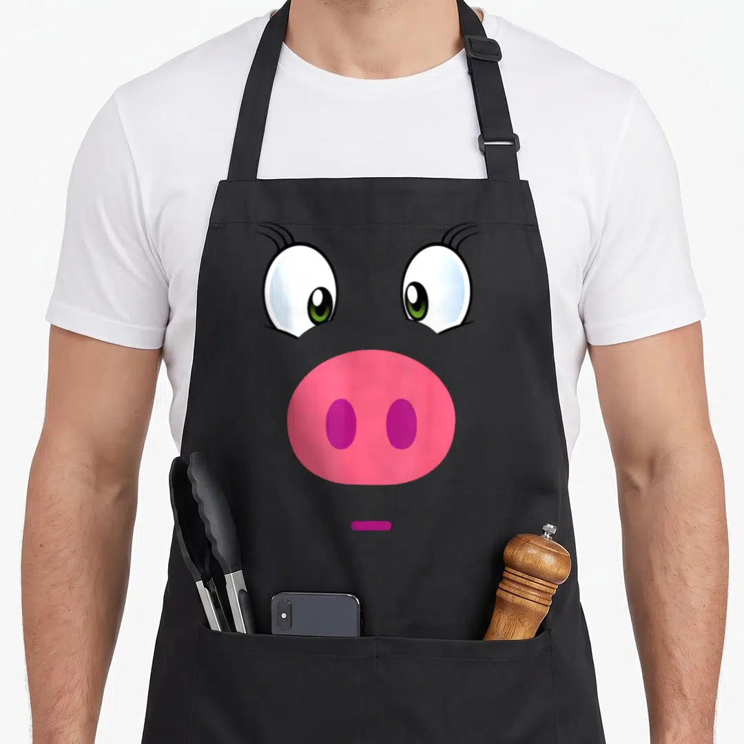 Piggy face apron - Cartoon pig face with pink snout, purple nostrils, green eyes, and small purple mouth line — shown on a black cotton canvas apron against a white background