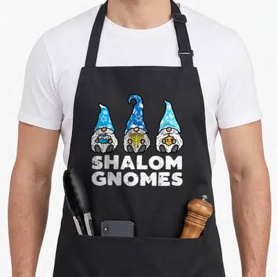 Shalom hanukkah apron - Three gnomes holding dreidel menorah and token below white Shalom Gnomes text — shown on a black cotton canvas apron against a white background
