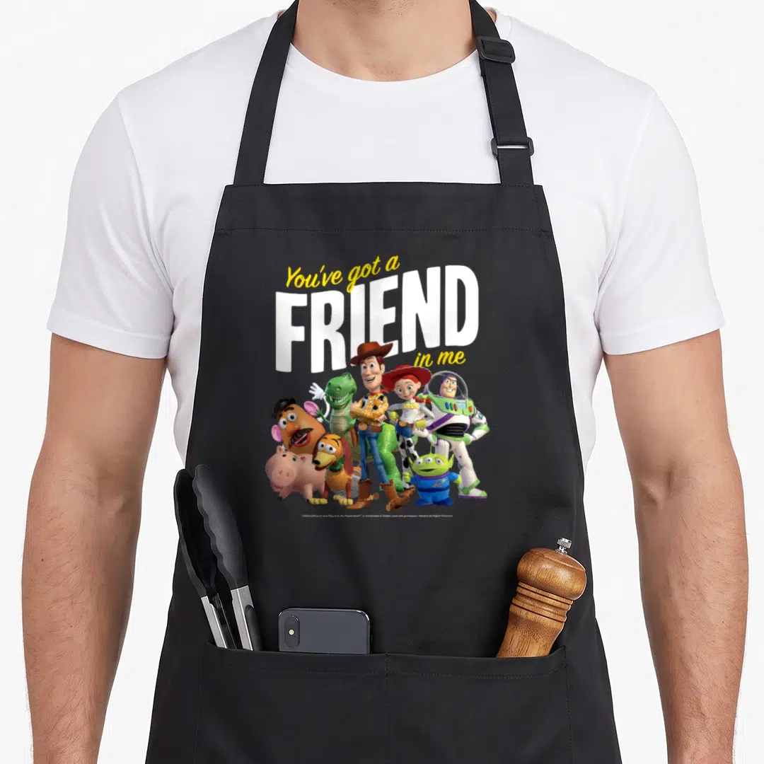 Cute friends apron - Toy Story characters cluster below You've Got a Friend in Me text in white, yellow, blue, green — shown on a black cotton canvas apron against a white background