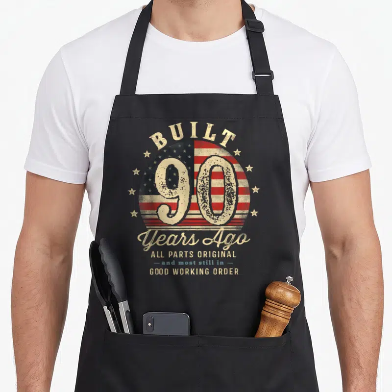 Flag birthday apron - Distressed USA flag circle with number 90 and text Built Years Ago Good Working Order — shown on a black cotton canvas apron against a white background