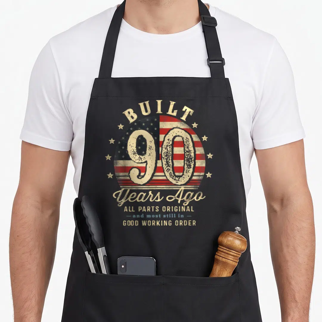 Flag birthday apron - Distressed USA flag circle with number 90 and text Built Years Ago Good Working Order — shown on a black cotton canvas apron against a white background