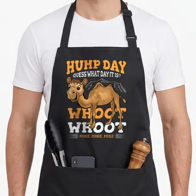 Wednesday camel apron - Cartoon camel with HUMP DAY text and WHOOT WHOOT MIKE stacked above and below — shown on a black cotton canvas apron against a white background