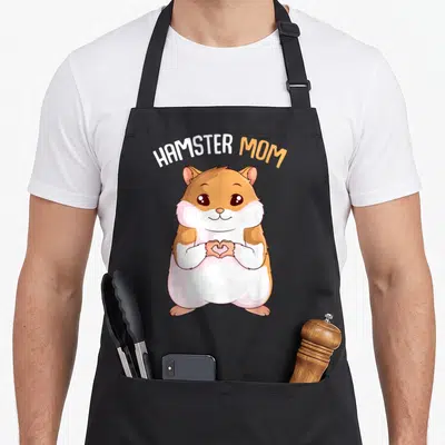 Hammy pet apron - Cartoon hamster making a heart shape with paws under arched HAMSTER MOM text in orange and white — shown on a black cotton canvas apron against a white background