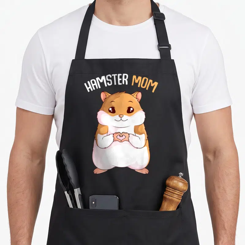 Hammy pet apron - Cartoon hamster making a heart shape with paws under arched HAMSTER MOM text in orange and white — shown on a black cotton canvas apron against a white background