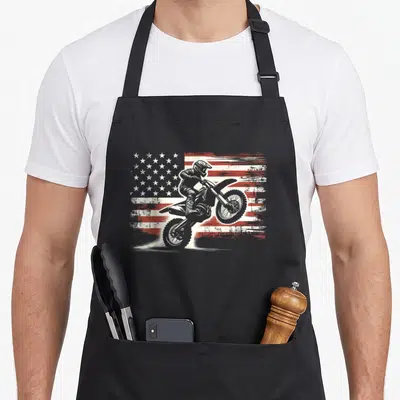 Biker american flag apron - Motocross rider on dirt bike in front of distressed red and white American flag graphic — shown on a black cotton canvas apron against a white background