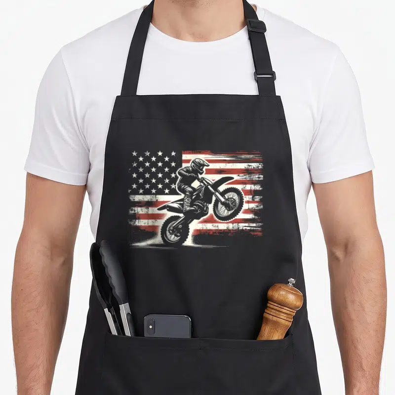 Biker american flag apron - Motocross rider on dirt bike in front of distressed red and white American flag graphic — shown on a black cotton canvas apron against a white background