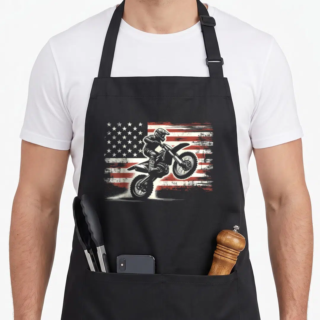 Biker american flag apron - Motocross rider on dirt bike in front of distressed red and white American flag graphic — shown on a black cotton canvas apron against a white background