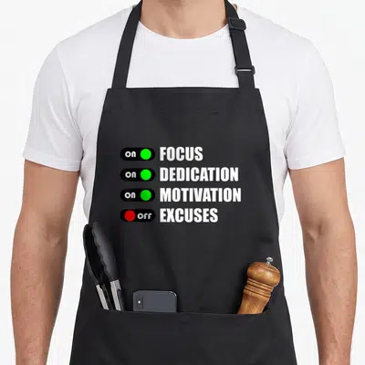 Focus dedication apron - Four toggle switches showing on FOCUS DEDICATION MOTIVATION and off EXCUSES in white black — shown on a black cotton canvas apron against a white background