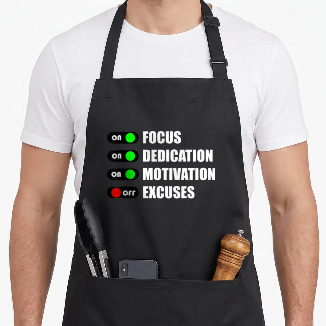 Focus dedication apron - Four toggle switches showing on FOCUS DEDICATION MOTIVATION and off EXCUSES in white black — shown on a black cotton canvas apron against a white background