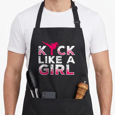 Kick kickboxing apron - Pink martial artist kicking above KICK LIKE A GIRL text in distressed white and pink — shown on a black cotton canvas apron against a white background