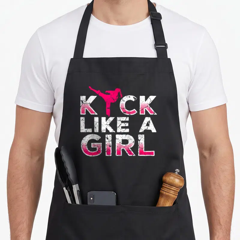 Kick kickboxing apron - Pink martial artist kicking above KICK LIKE A GIRL text in distressed white and pink — shown on a black cotton canvas apron against a white background