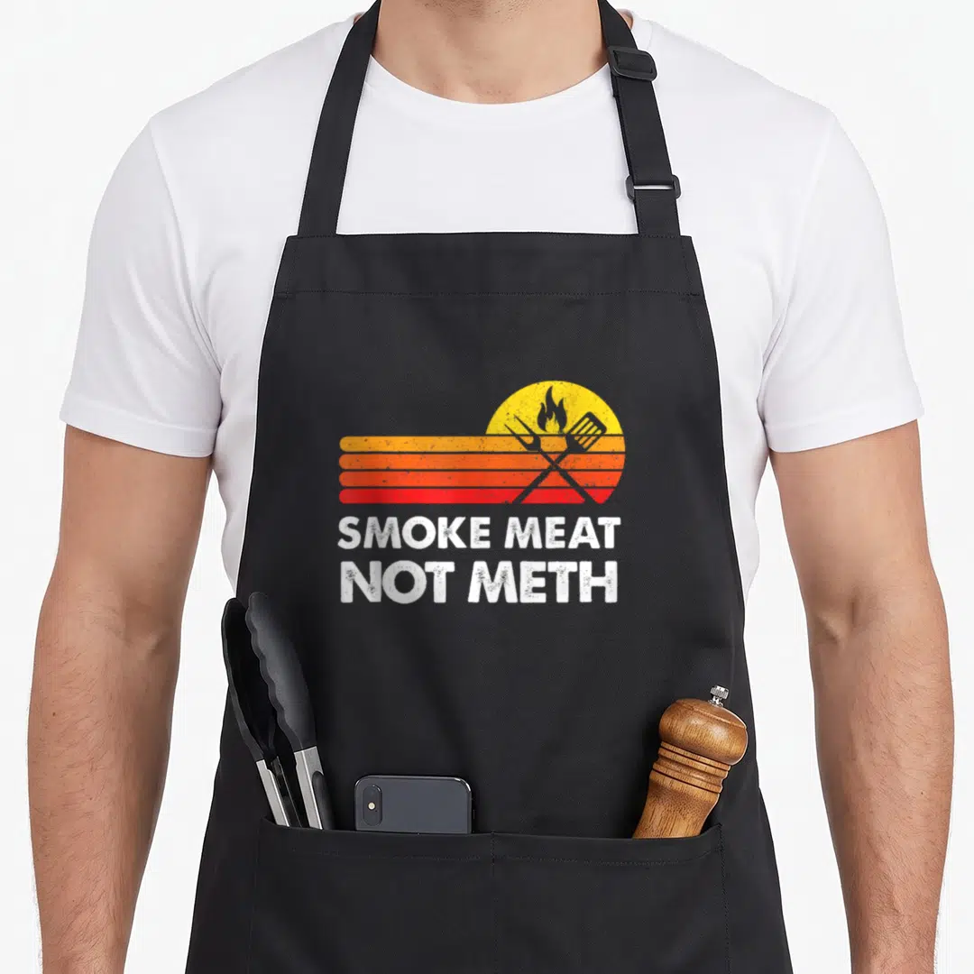Brisket meat aprons - Retro yellow sun with crossed grill tools and orange red stripes above white text reading Smoke — shown on a black cotton canvas apron against a white background