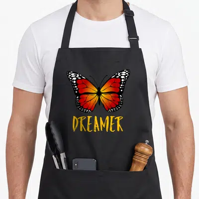 Save butterfly apron - Realistic monarch butterfly with orange wings above yellow DREAMER text — shown on a black cotton canvas apron against a white background