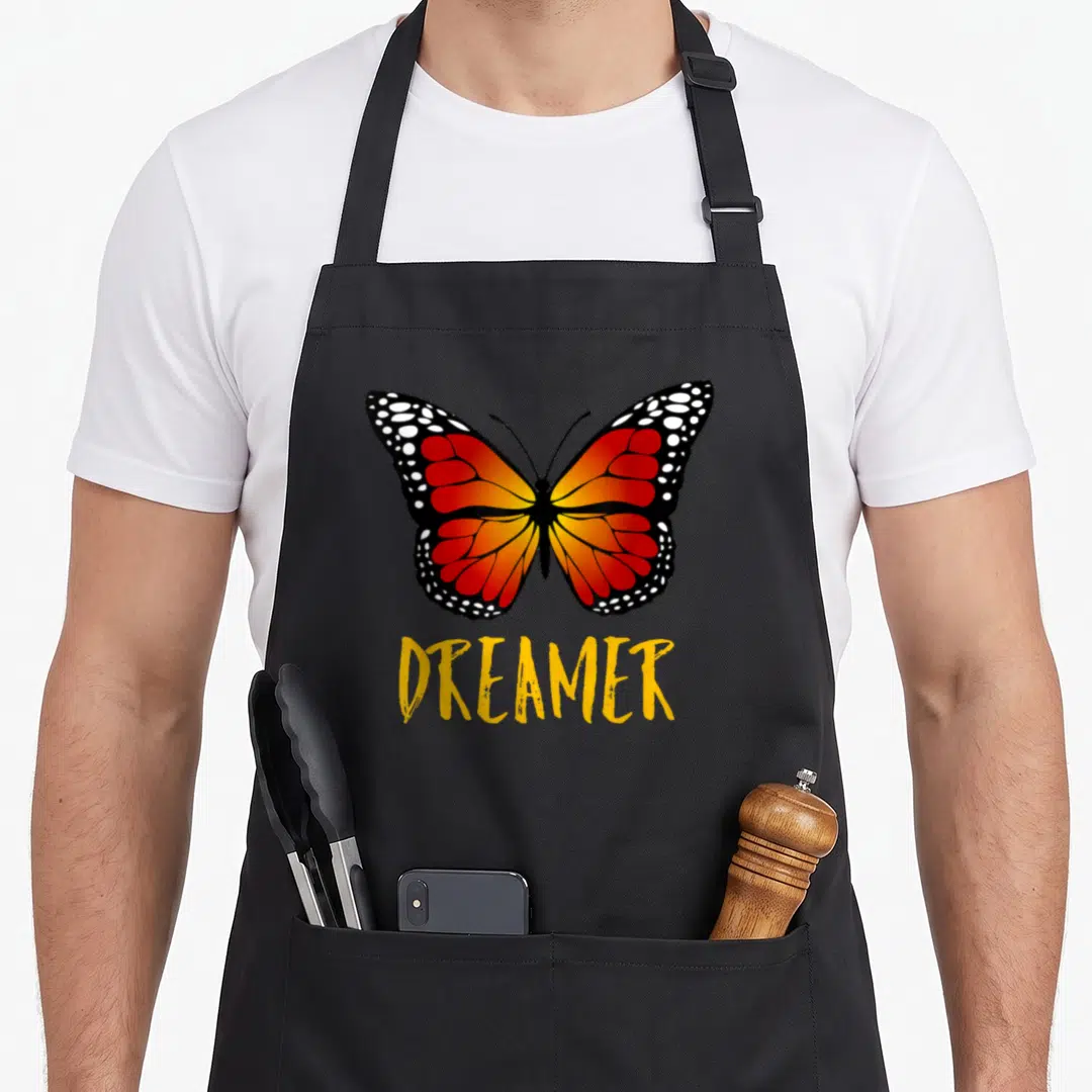 Save butterfly apron - Realistic monarch butterfly with orange wings above yellow DREAMER text — shown on a black cotton canvas apron against a white background
