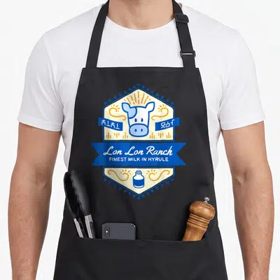 Lon milk aprons - Cartoon cow head in cream hexagon with blue ribbon reading Lon Lon Ranch and milk bottle — shown on a black cotton canvas apron against a white background