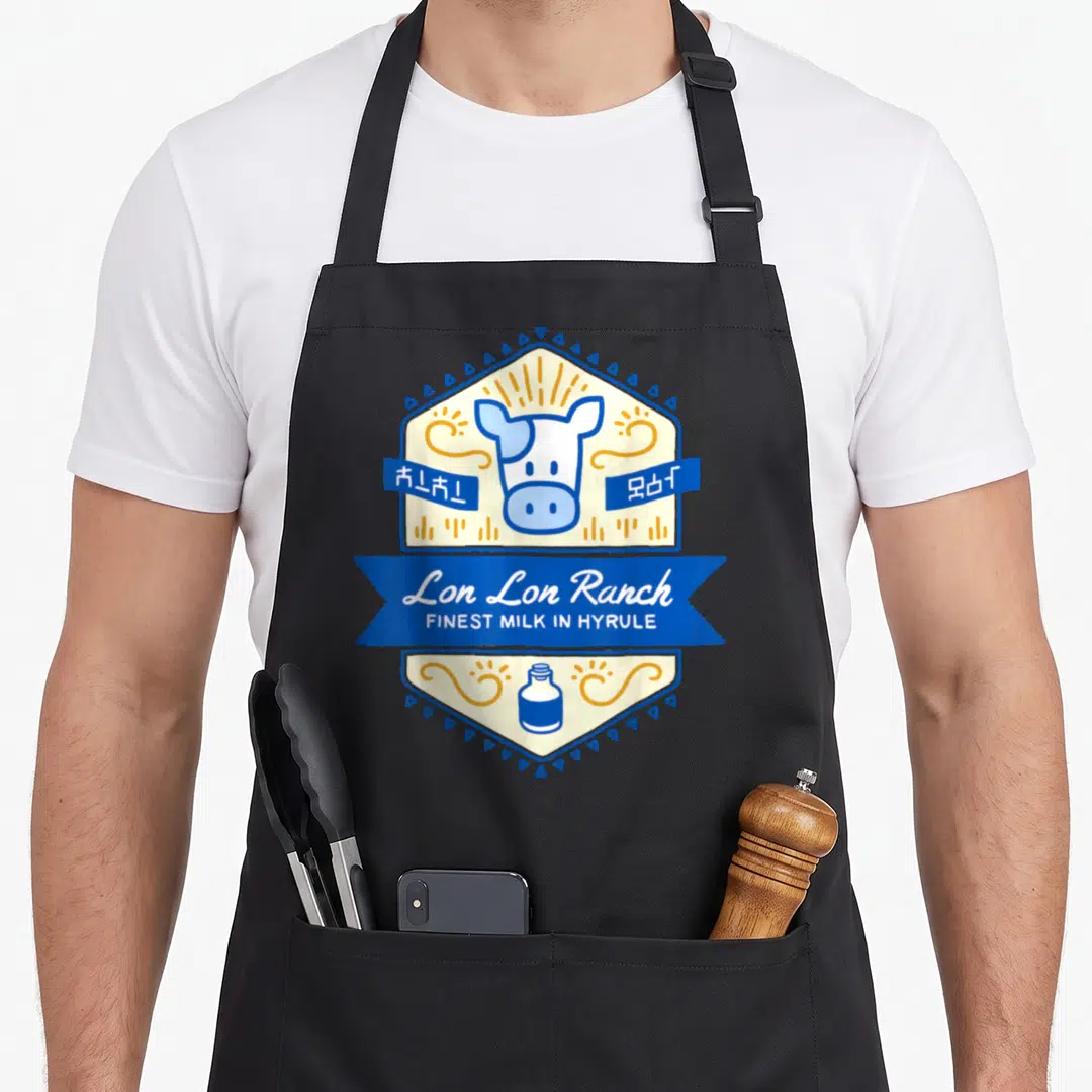 Lon milk aprons - Cartoon cow head in cream hexagon with blue ribbon reading Lon Lon Ranch and milk bottle — shown on a black cotton canvas apron against a white background