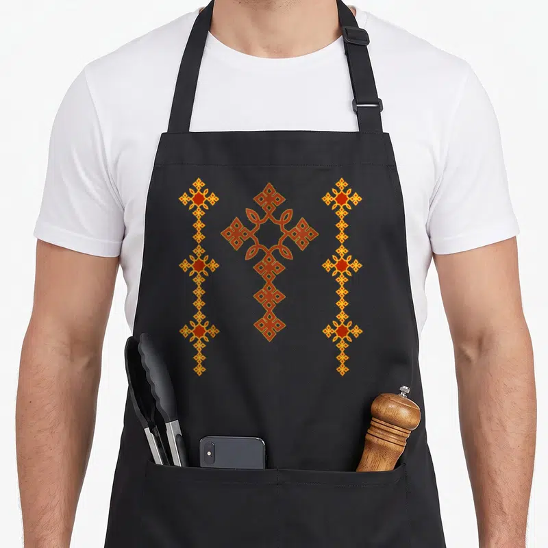 Traditional cross apron - Three vertical orange and red geometric columns with central interlaced cross design — shown on a black cotton canvas apron against a white background