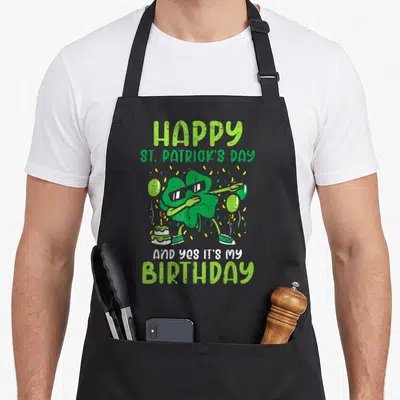 Bday st patrick day aprons - Cartoon shamrock dabbing with sunglasses near cake and balloons with birthday text — shown on a black cotton canvas apron against a white background