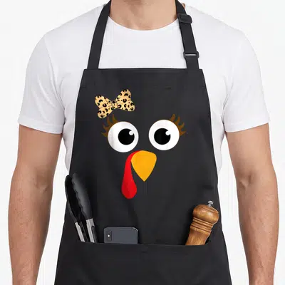Turkey leopard apron - Cartoon turkey face with leopard bow, orange beak, red wattle, and big eyes — shown on a black cotton canvas apron against a white background