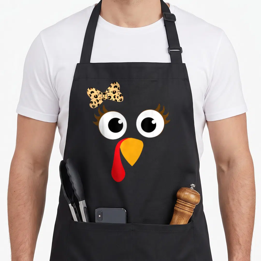 Turkey leopard apron - Cartoon turkey face with leopard bow, orange beak, red wattle, and big eyes — shown on a black cotton canvas apron against a white background