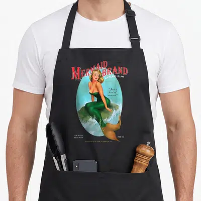 Jamaican mermaid apron - Retro mermaid label with red text, green tail, and Just a hint of seaweed quote inside an oval — shown on a black cotton canvas apron against a white background
