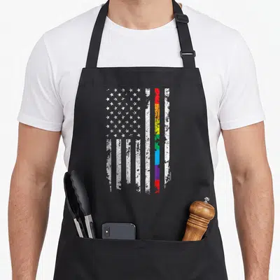 Gay line apron - Vertical distressed American flag with central rainbow stripe in white red orange yellow green blue — shown on a black cotton canvas apron against a white background