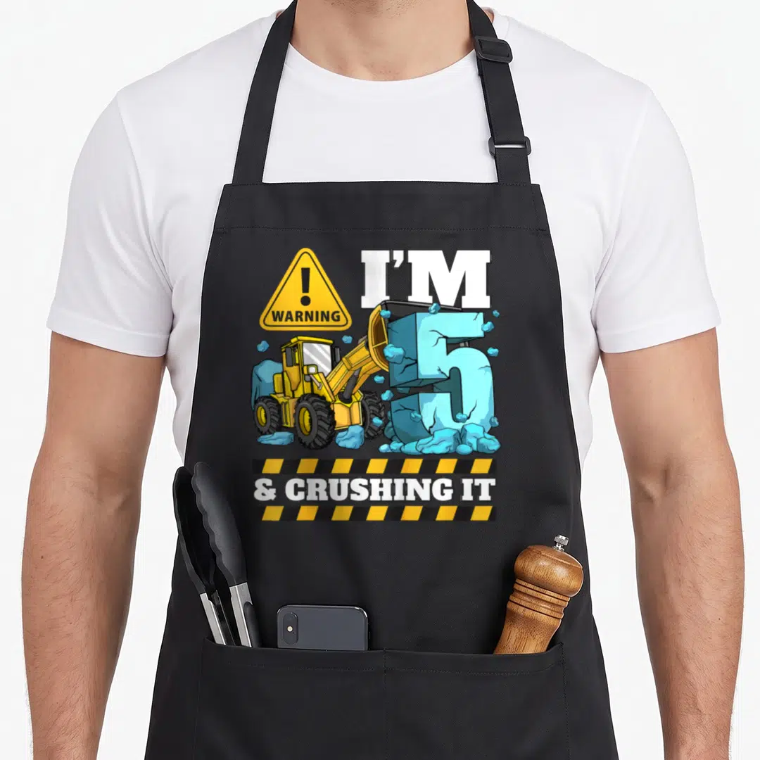 Construction 5th birthday truck apron - Yellow front loader crushing blue number 5 with warning sign and hazard stripes — shown on a black cotton canvas apron against a white background