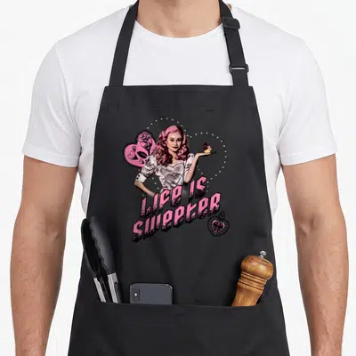 Sweeter novelty apron - Woman holding cupcake with LIFE IS SWEETER text and pink heart arch — shown on a black cotton canvas apron against a white background