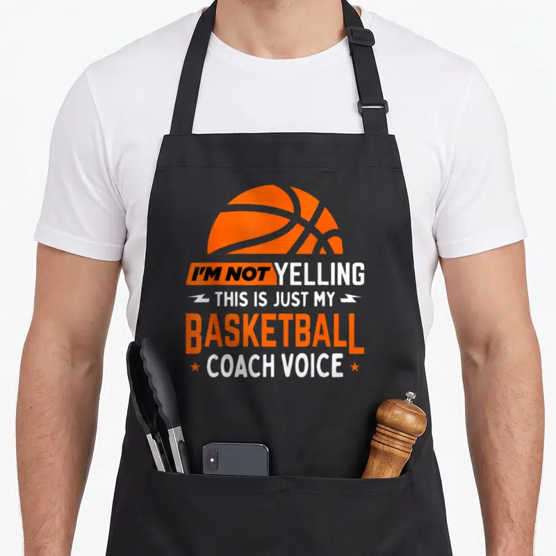 Yelling coaching apron - Orange basketball graphic above stacked I'm Not Yelling Basketball Coach Voice text — shown on a black cotton canvas apron against a white background