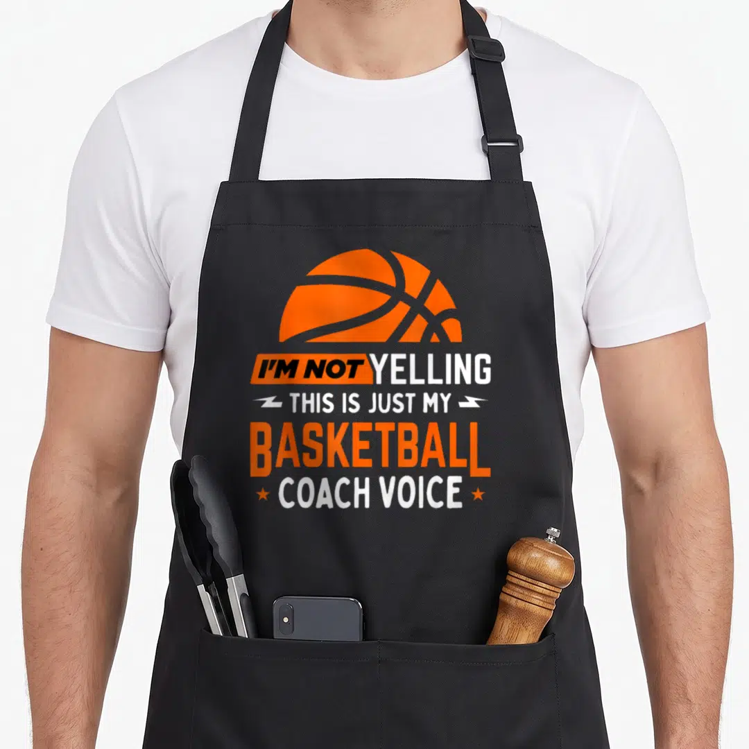 Yelling coaching apron - Orange basketball graphic above stacked I'm Not Yelling Basketball Coach Voice text — shown on a black cotton canvas apron against a white background