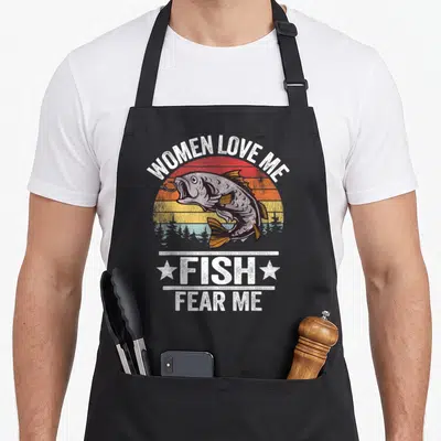 Fear fisher fish apron - Retro sunset graphic with jumping fish and text Women Love Me Fish Fear Me — shown on a black cotton canvas apron against a white background