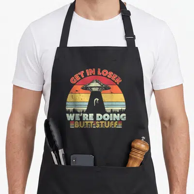 Loser funny soul apron - Retro UFO abduction scene with GET IN LOSER WE'RE DOING BUTT STUFF text over striped sun — shown on a black cotton canvas apron against a white background