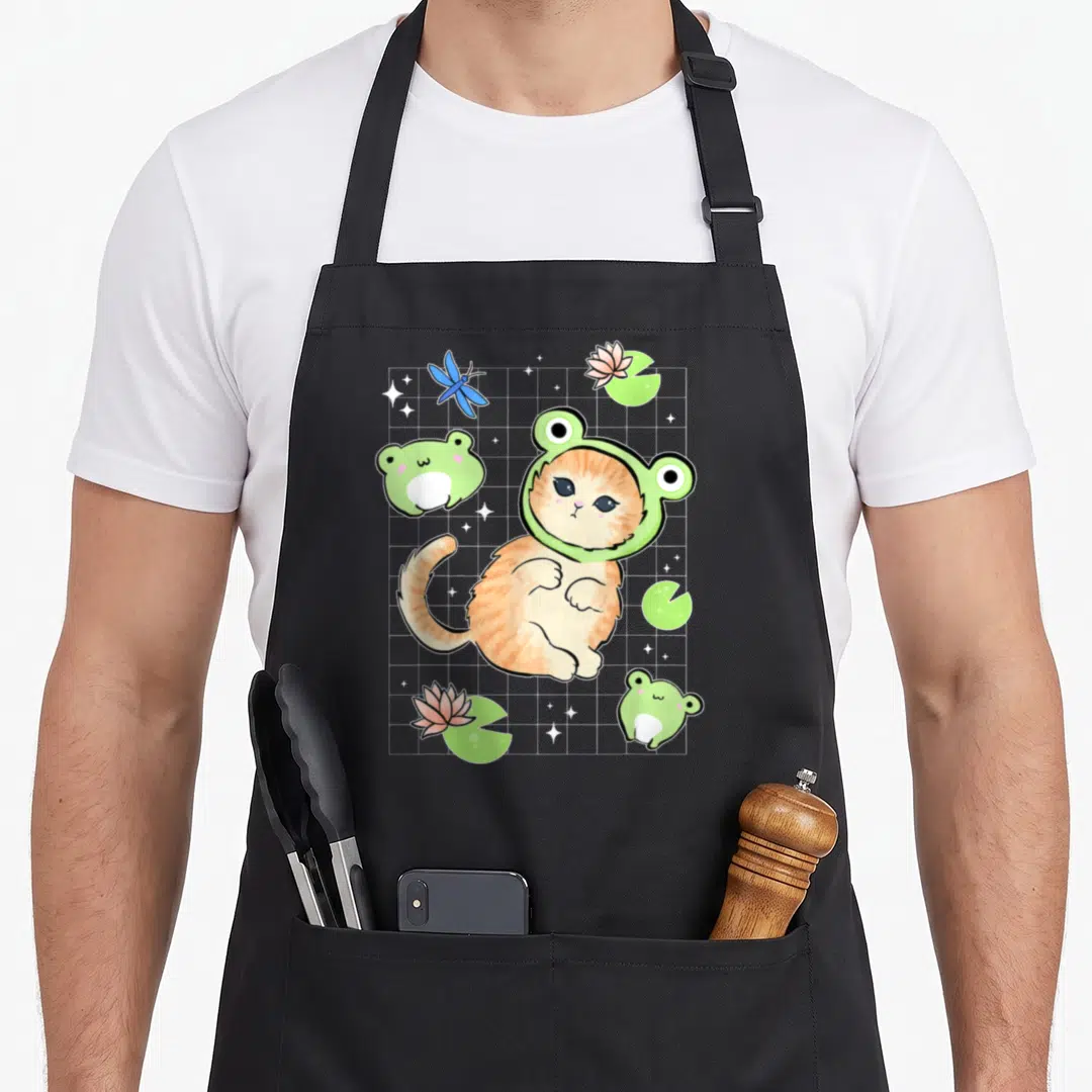 Frog cat cottagecore apron - Orange cat in green frog hat with lily pads and blue dragonfly on white grid — shown on a black cotton canvas apron against a white background