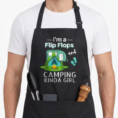 Flip camping apron - Text I'm a Flip Flops and CAMPING KINDA GIRL around a blue camper, tent, and orange fire — shown on a black cotton canvas apron against a white background
