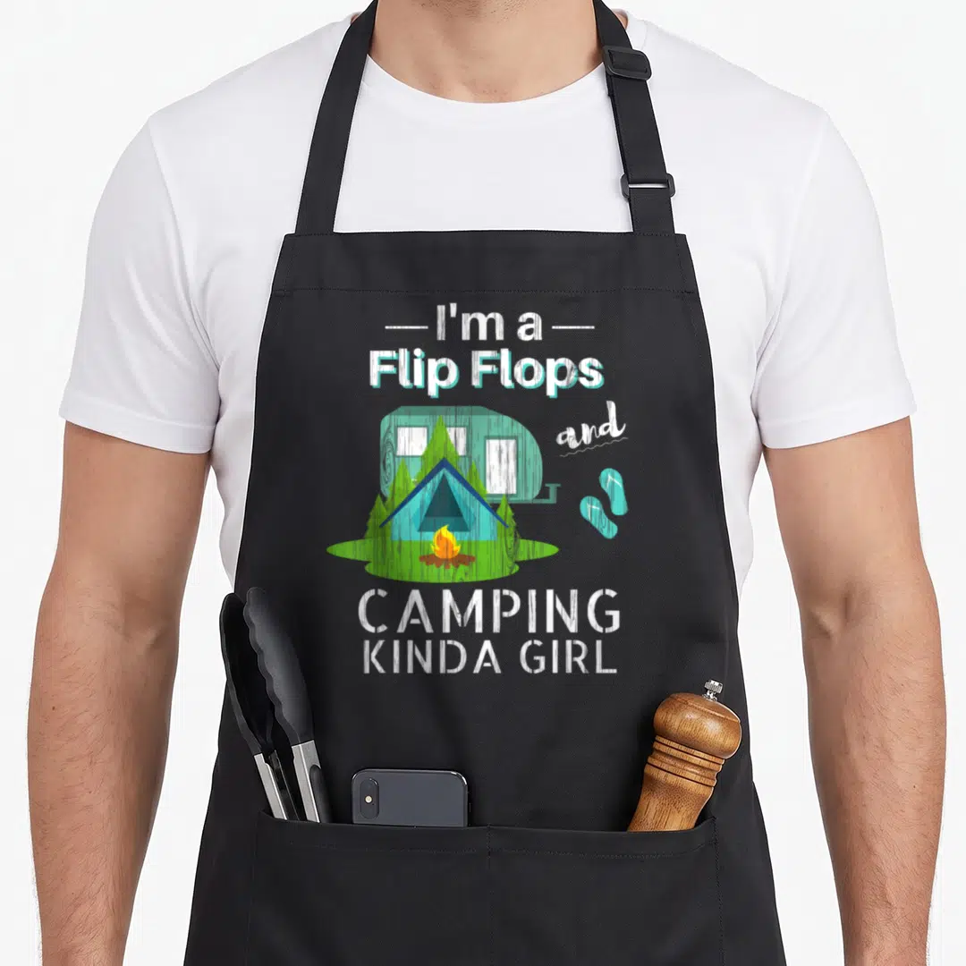 Flip camping apron - Text I'm a Flip Flops and CAMPING KINDA GIRL around a blue camper, tent, and orange fire — shown on a black cotton canvas apron against a white background