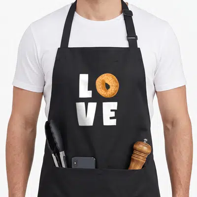 Bagel themed aprons - White block letters LVE with golden brown bagel replacing O — shown on a black cotton canvas apron against a white background