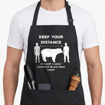 Distance bear apron - White bear silhouette between two people with distance measurement arrow and text — shown on a black cotton canvas apron against a white background