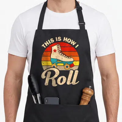 Speed roller skating apron - Retro quad skate with striped sunset circle and arched text reading THIS IS HOW I Roll — shown on a black cotton canvas apron against a white background