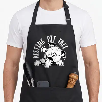 Resting dog face apron - Cartoon dog resting with tongue out, arched text RESTING PIT FACE, bone, and paw print — shown on a black cotton canvas apron against a white background