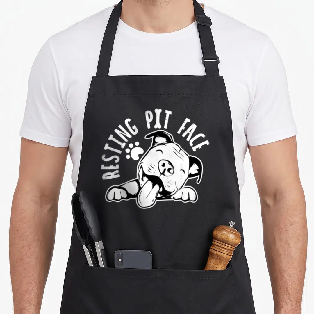 Resting dog face apron - Cartoon dog resting with tongue out, arched text RESTING PIT FACE, bone, and paw print — shown on a black cotton canvas apron against a white background