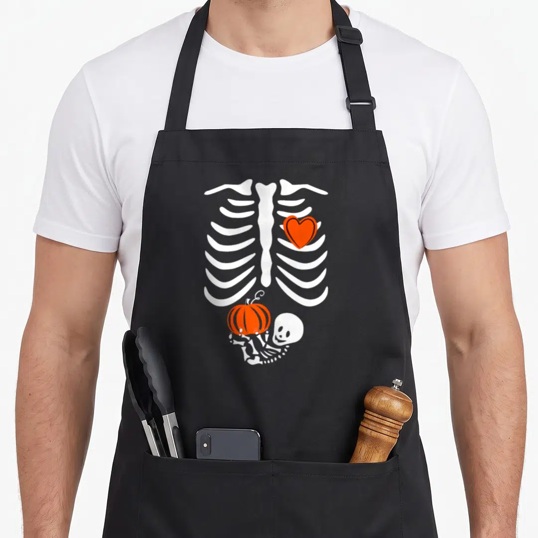 Pregnant halloween apron - White skeleton ribcage with orange heart and small skeleton lying holding orange pumpkin — shown on a black cotton canvas apron against a white background