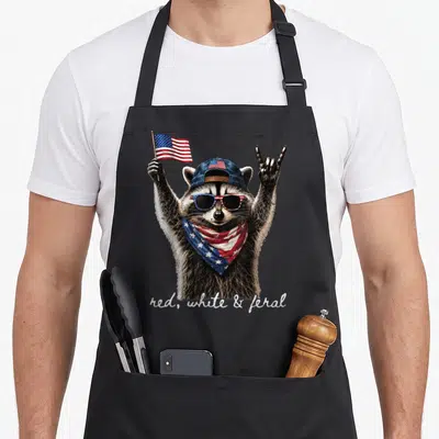 Feral 4th of july apron - Realistic raccoon with sunglasses, bandana, and flag making a rock sign with red white — shown on a black cotton canvas apron against a white background