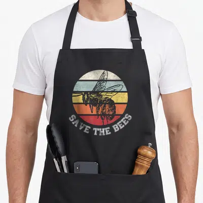 Distressed bee apron - Retro striped circle with bee illustration and arched SAVE THE BEES text — shown on a black cotton canvas apron against a white background