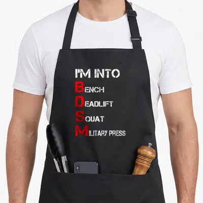 Bdsm bench apron - Stacked white and red text listing gym exercises with vertical BDSM acronym — shown on a black cotton canvas apron against a white background
