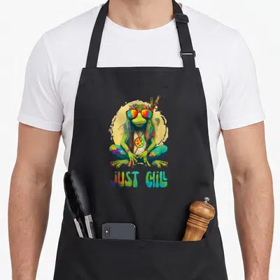 Chill frog aprons - Green frog with rainbow glasses and JUST CHILL text — shown on a black cotton canvas apron against a white background