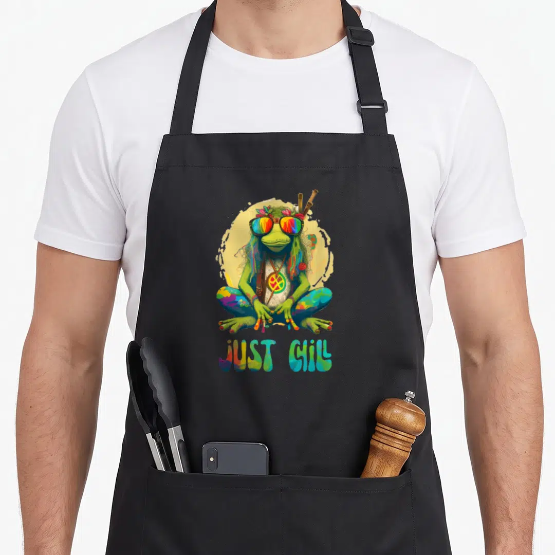 Chill frog aprons - Green frog with rainbow glasses and JUST CHILL text — shown on a black cotton canvas apron against a white background